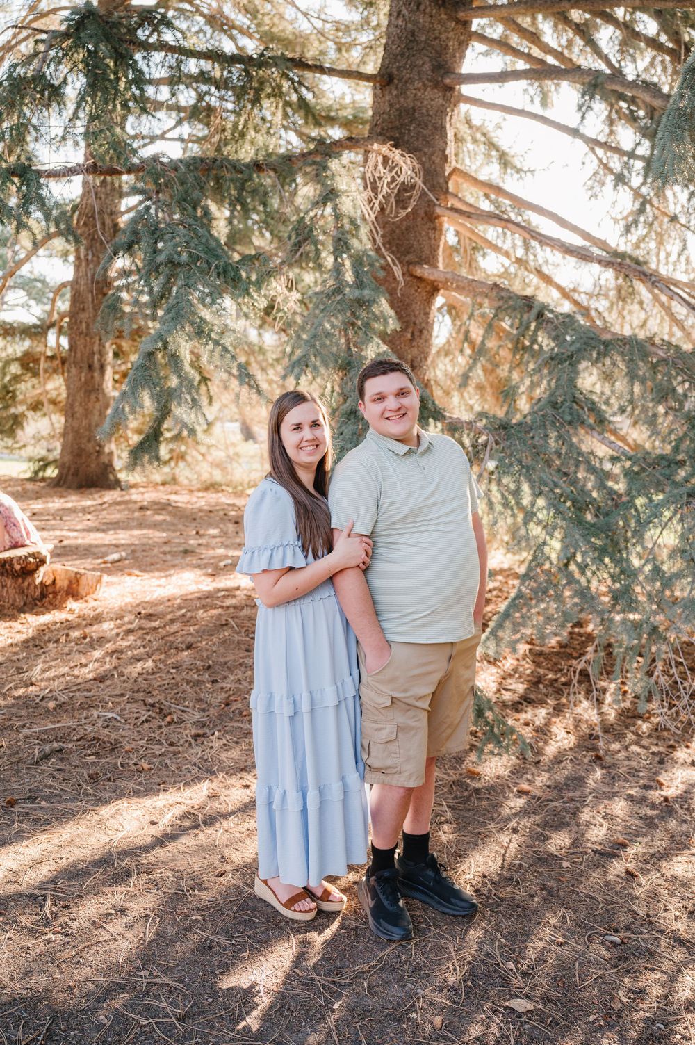A young couple smiles together outdoors under pine trees, woman in blue maxi dress, man in khaki shorts and green polo.