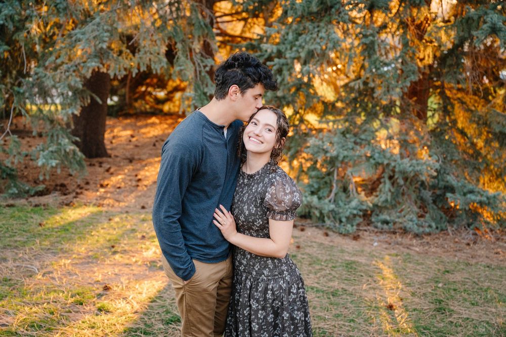 Couple sharing a tender moment outdoors at golden hour beneath evergreen trees in a romantic engagement photo.