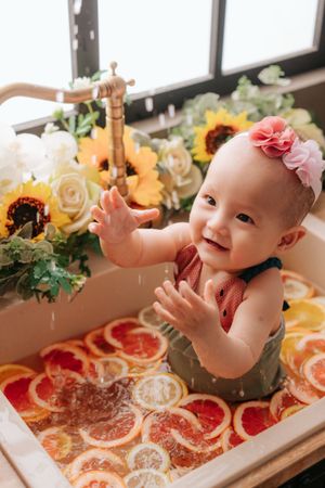 Smiling baby girl in floral headband sitting in a sink filled with citrus slices and water, surrounded by sunflowers.