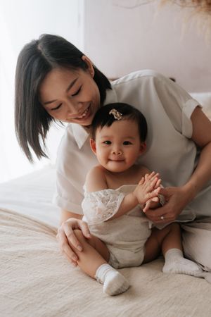 Smiling Asian mother sitting on bed with happy baby girl in white lace outfit and hair clip.