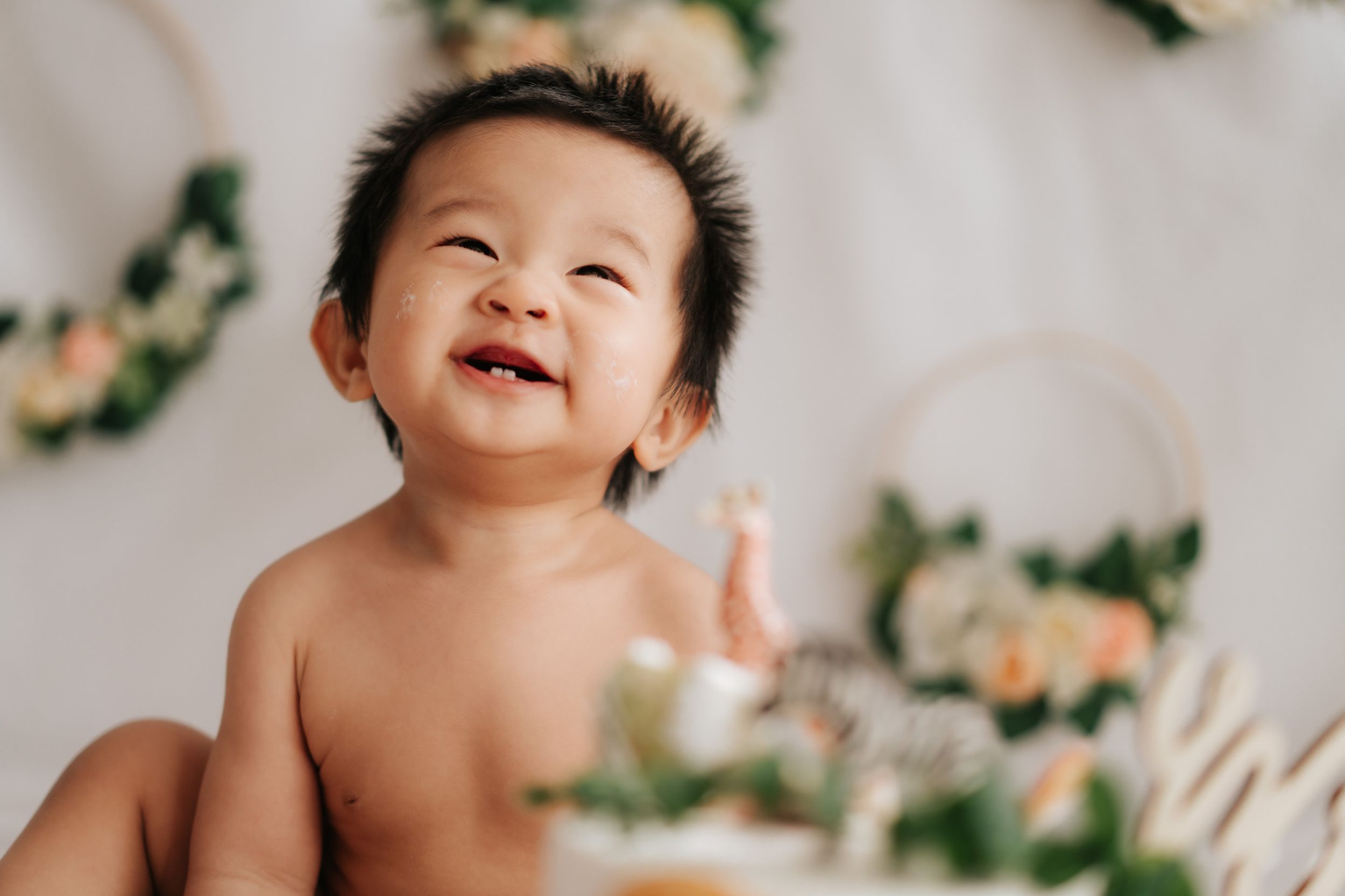 Happy Asian baby smiling joyfully, sitting shirtless surrounded by floral decorations and greenery wreaths.
