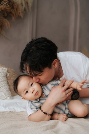 A loving father gently kisses his baby on the head while holding them on a cozy bed.