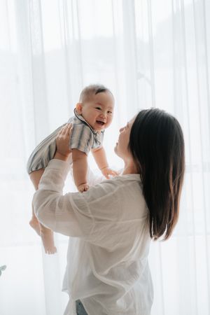 A joyful mother lifts her laughing baby in the air near a bright white curtained window.