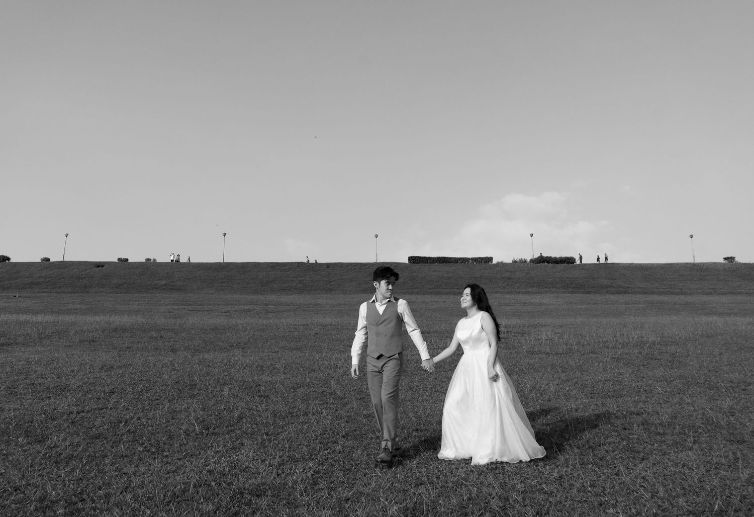 A couple in wedding attire share a playful moment together on a lawn with white buildings in the background.