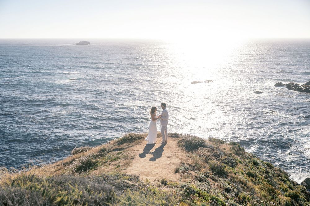 Couple standing on a coastal cliff overlooking the Pacific Ocean at sunset in a romantic engagement photo.