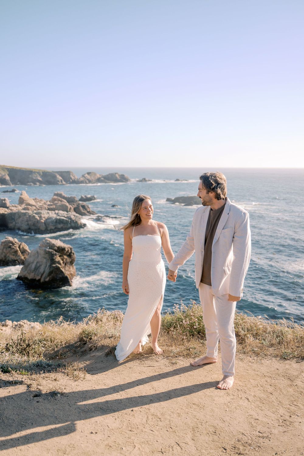 A couple in wedding attire holds hands on a coastal cliff with rocky ocean views under a clear blue sky.