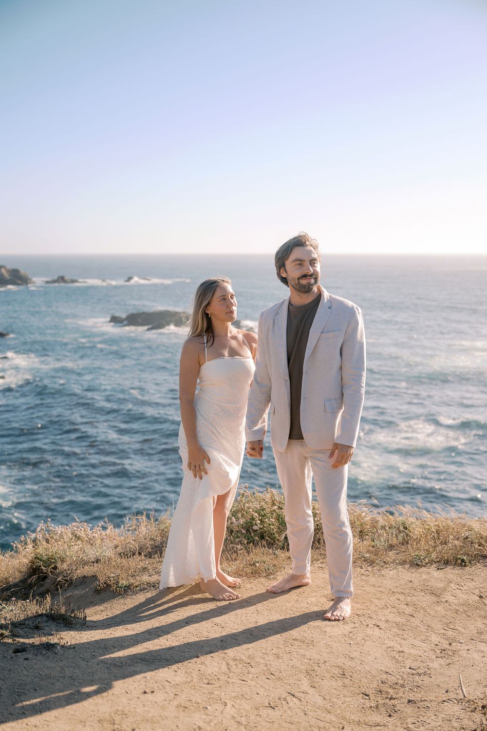 Couple holding hands on coastal cliff overlooking blue ocean, woman in white dress, man in light suit, sunny day.