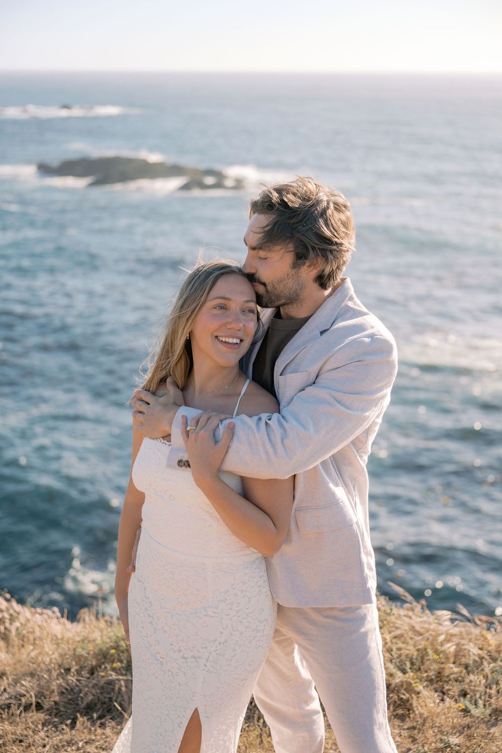 Couple embracing on coastal cliff, woman in white lace dress smiling, man in beige suit, ocean backdrop.