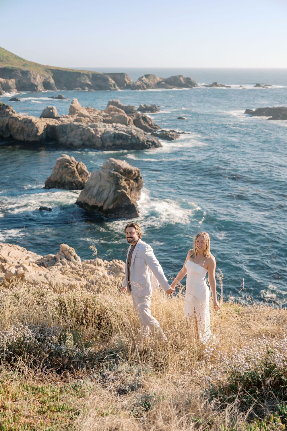 Couple in white holding hands on rocky Big Sur coastline with turquoise ocean waves and cliffs in background.