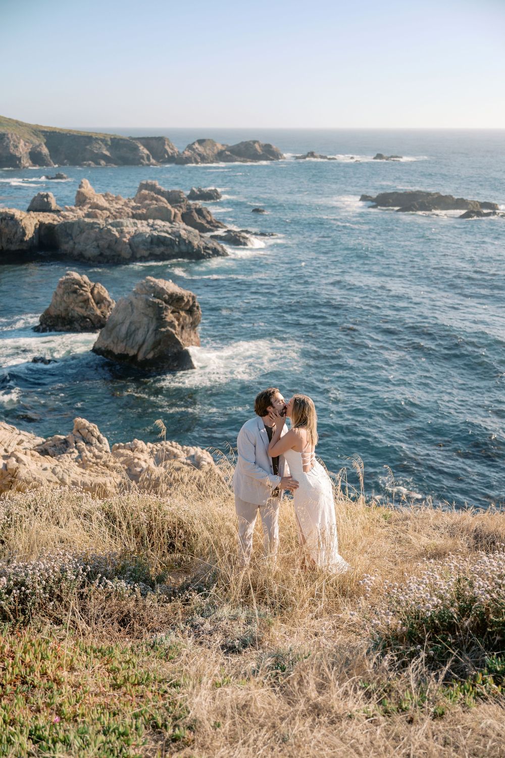 Couple kissing on rocky coastal cliffside in Big Sur, woman in white dress, man in light suit, blue ocean backdrop.