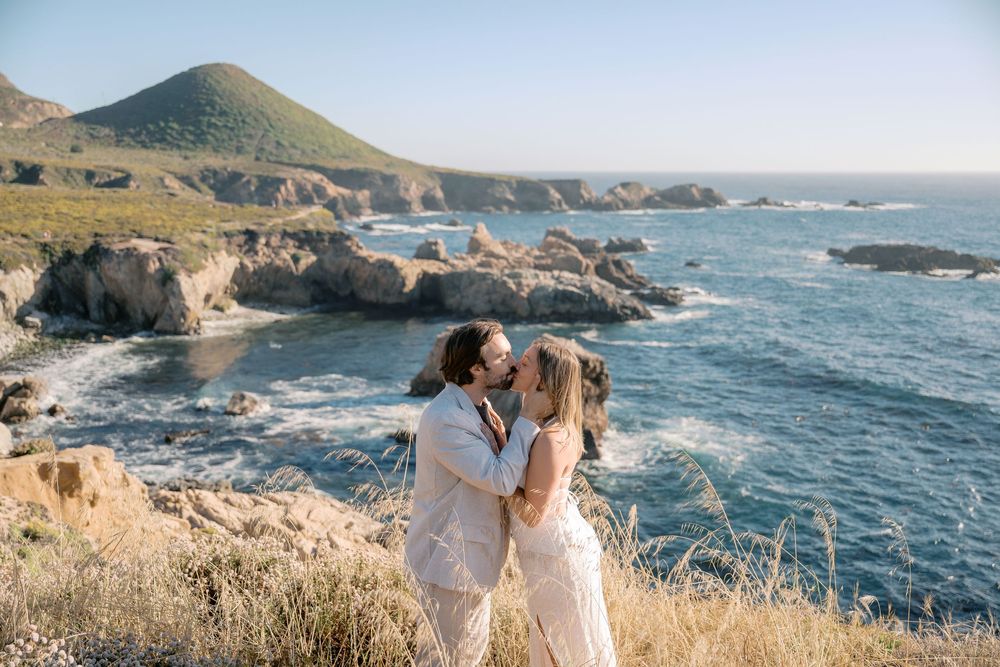 Couple sharing a romantic kiss on a coastal cliff overlooking the rocky Big Sur coastline and blue Pacific Ocean.