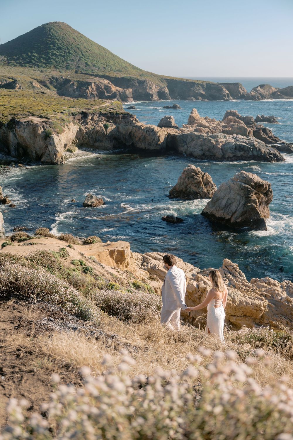 Couple in white holding hands on rocky Big Sur coastline with turquoise ocean and green hills.