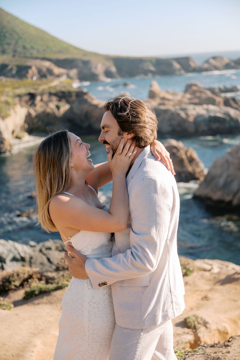 Couple in wedding attire embracing and laughing on rocky California coastal cliffs with turquoise ocean views.