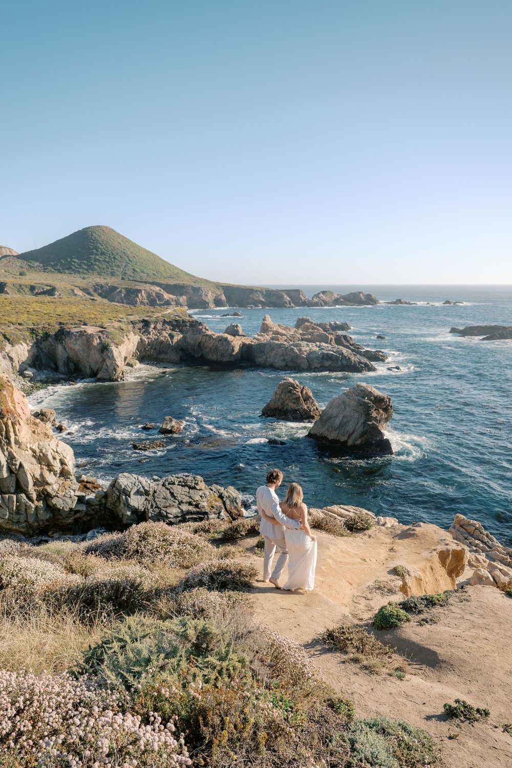 Couple in white attire standing on rocky coastal cliffs overlooking turquoise ocean waters at Big Sur, California.