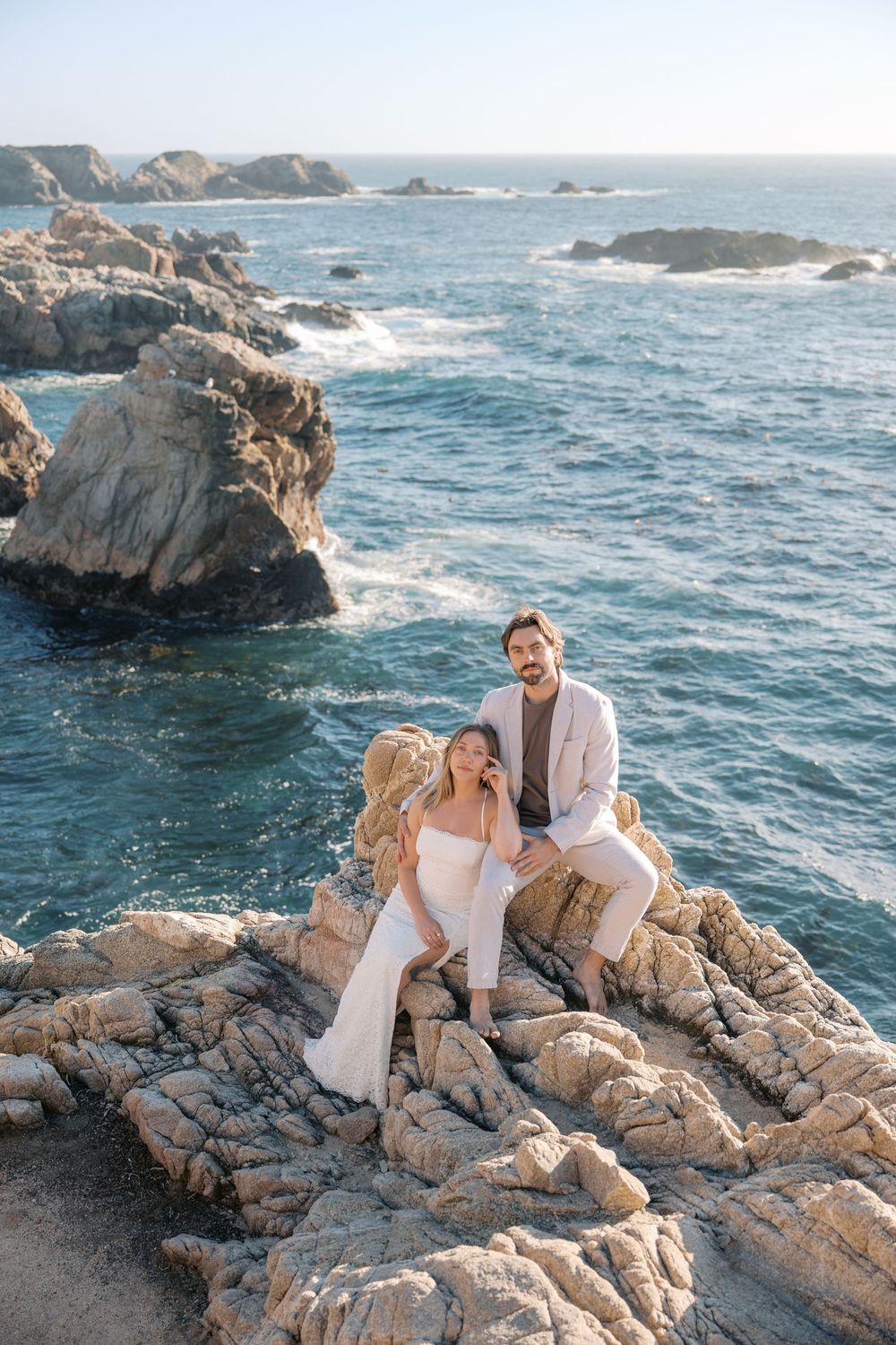 Romantic couple sitting together on rugged ocean rocks with crashing waves and blue sea in the background.