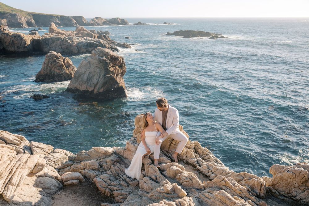 Couple in wedding attire sitting on coastal rocks with dramatic ocean waves and sea stacks in the background.