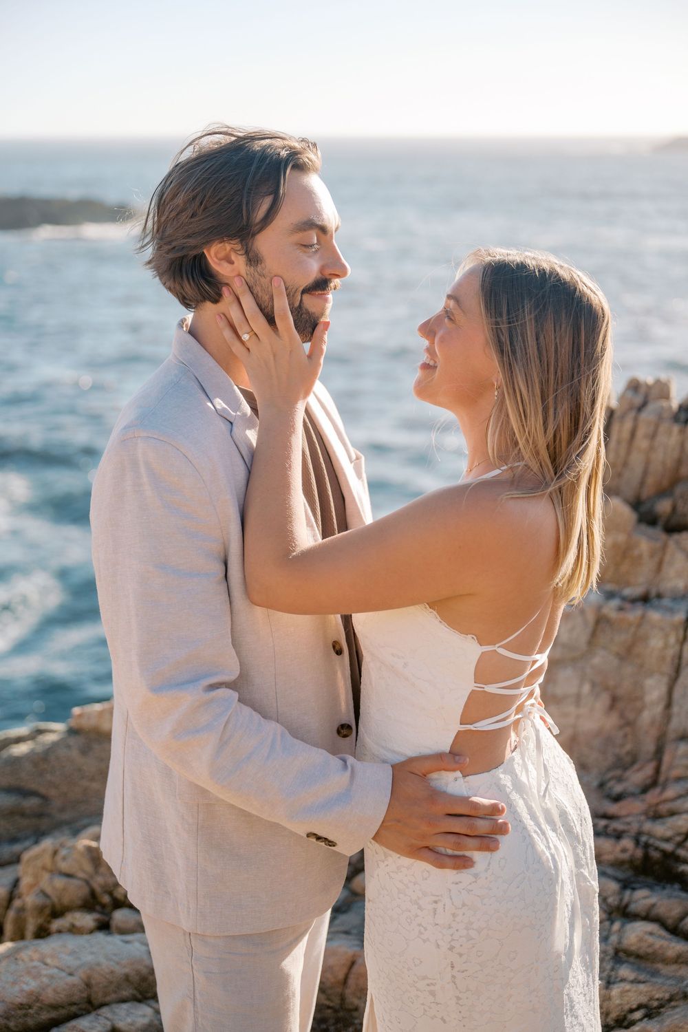 Smiling couple in elegant attire share a tender moment on ocean rocks, woman in white dress touches mans face.