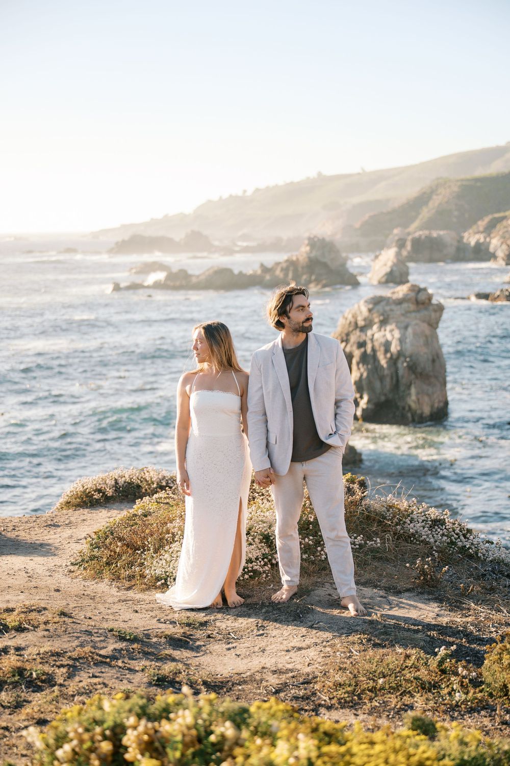 Couple in white dress and gray suit standing on coastal cliffs with ocean and rocks behind.