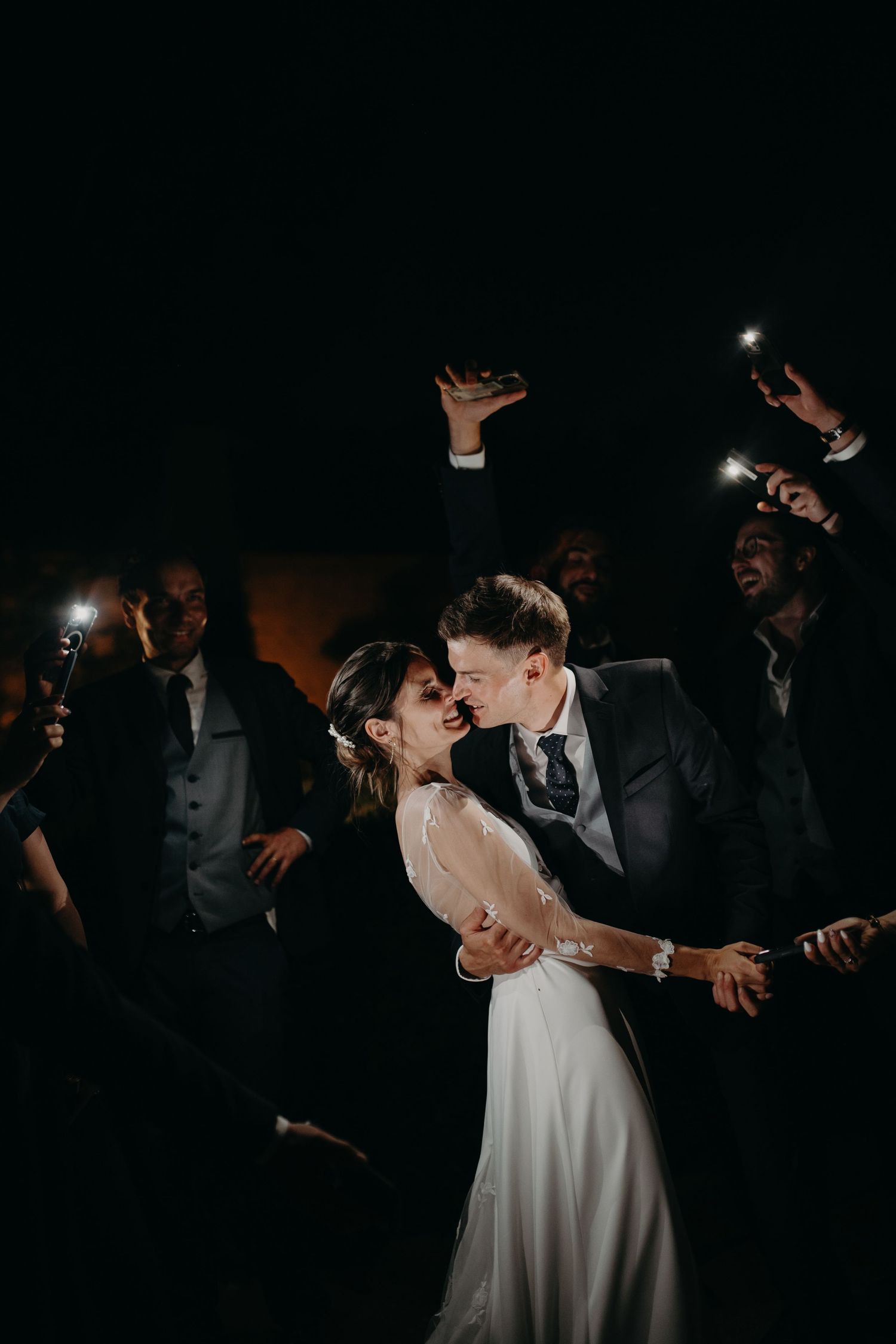 Black and white wedding photo of bride and groom kissing outdoors, with blurred guests and elegant chateau in background.