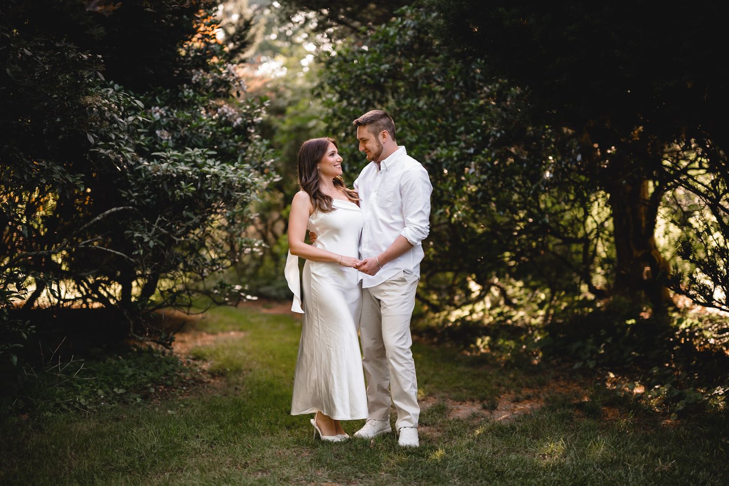 Couple in white attire standing together in a lush green garden, sharing a tender moment outdoors.