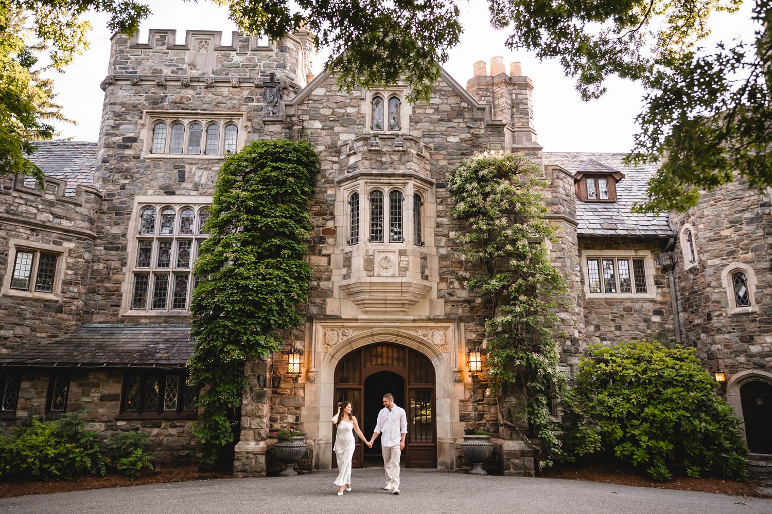 Couple holding hands in front of an ivy-covered stone castle wedding venue with arched entrance and lanterns.