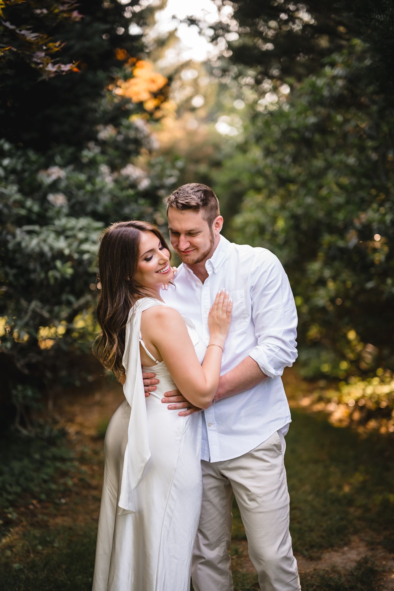 Couple embracing in lush garden, woman in white dress smiling, man in white shirt and khaki pants.