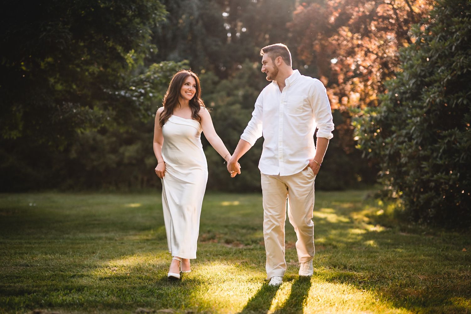 A couple holds hands and walks through a sunlit park, smiling at each other in matching white outfits.