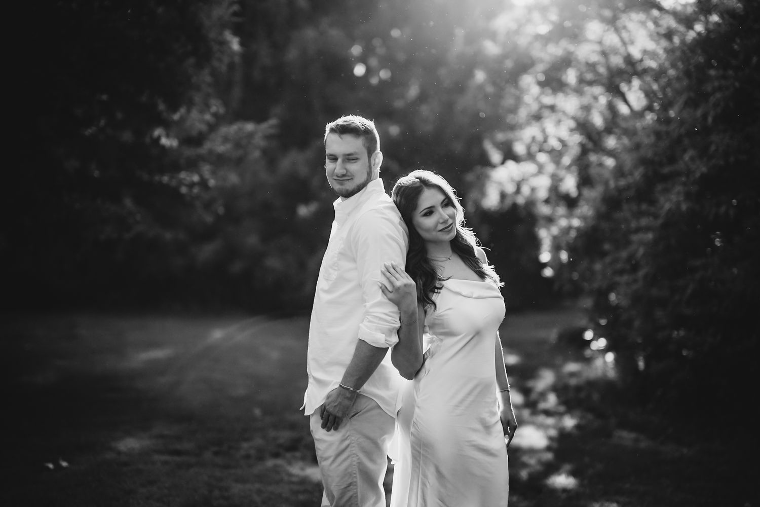 Black and white photo of a couple in white outfits embracing outdoors in a lush garden setting.
