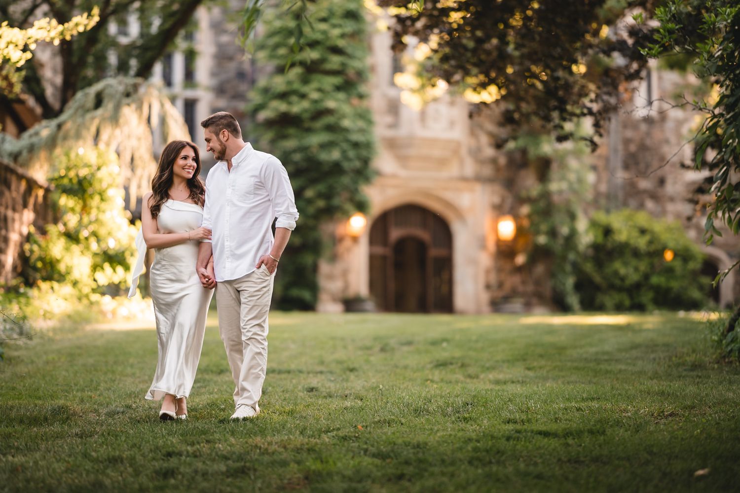 Couple walking hand-in-hand on green lawn before a stone building at golden hour.