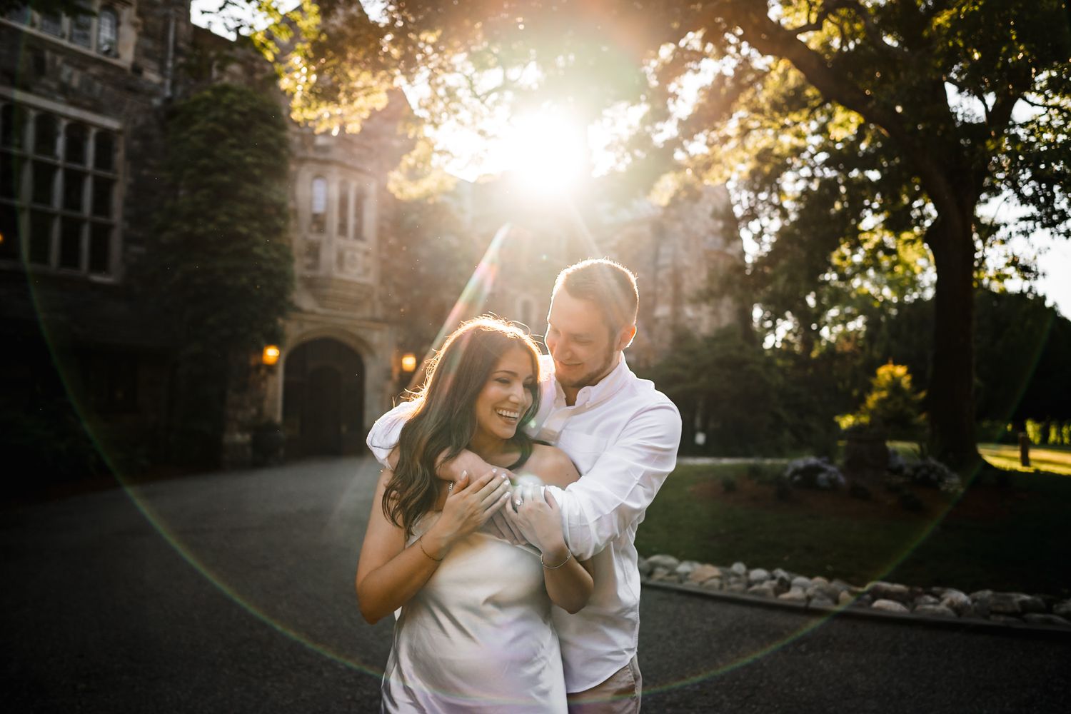 Couple embracing at golden hour outside a historic castle, bathed in warm sunlight filtering through trees.