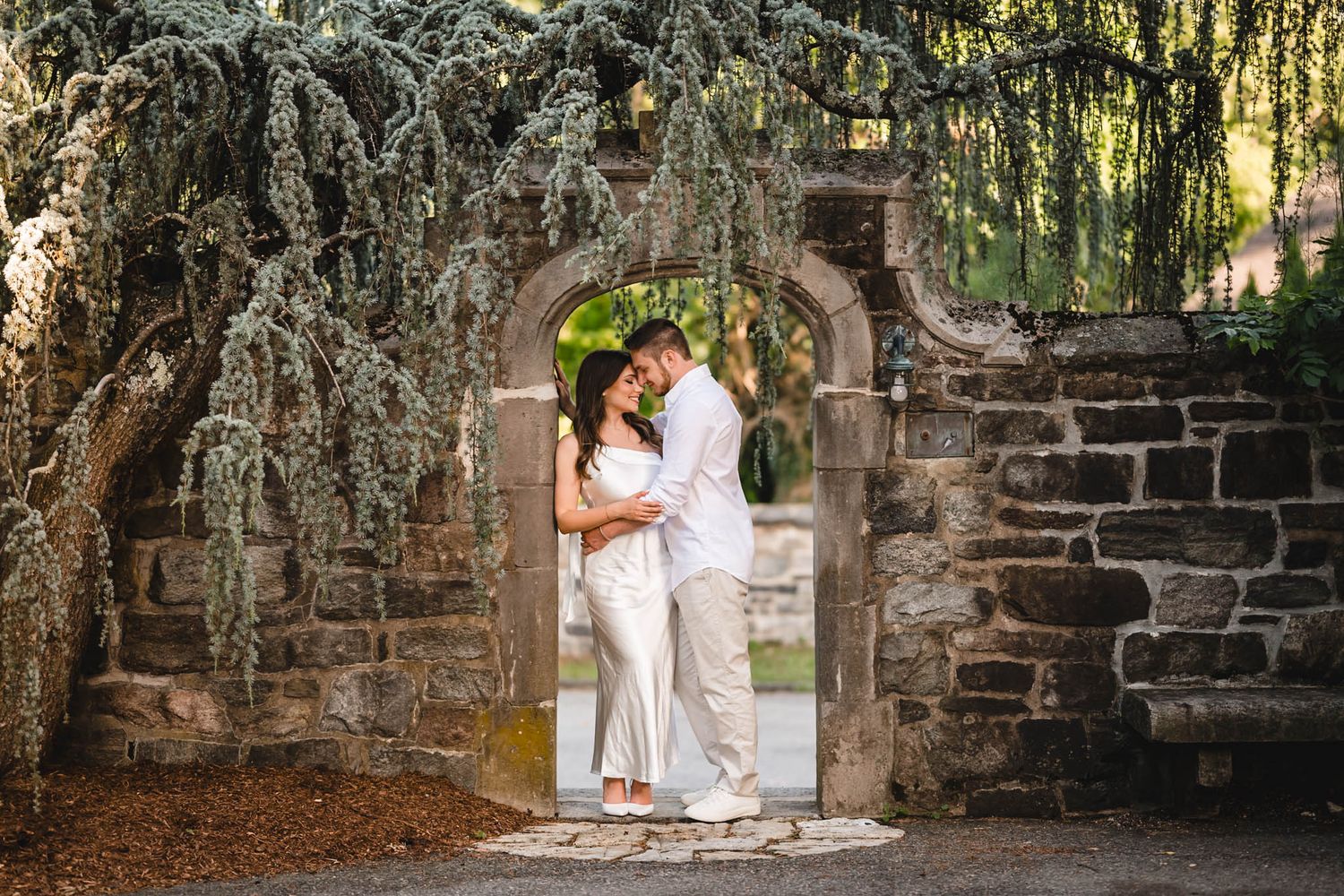 Couple embracing under a stone archway surrounded by lush weeping trees in a romantic outdoor garden setting.