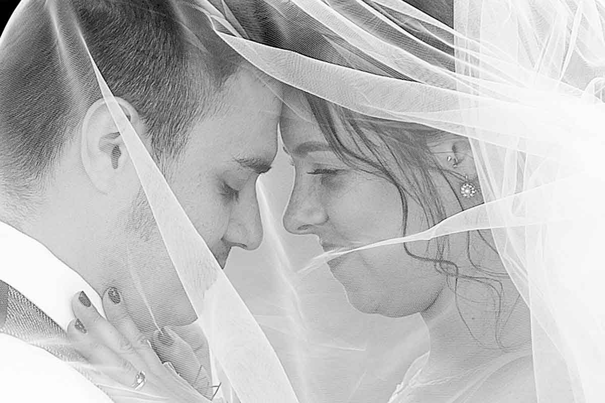 Black and white wedding photo of bride and groom touching foreheads under a flowing veil.