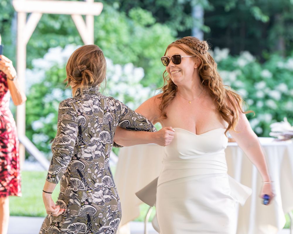 Bride in white strapless gown dancing with guest in floral dress at outdoor wedding reception.