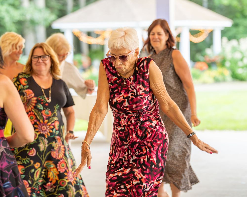 Elderly woman in pink floral dress dancing joyfully at an outdoor wedding reception with guests in background.