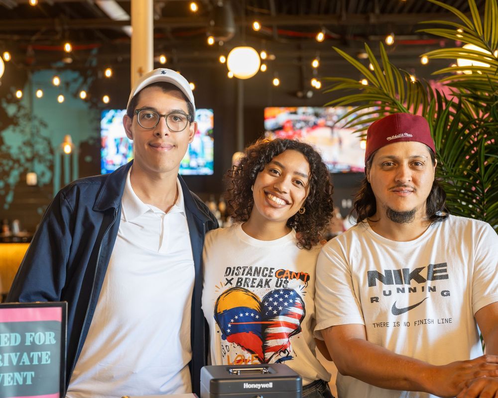 Three friends smiling together at a restaurant bar, wearing casual clothes including a Nike shirt and patriotic tee.
