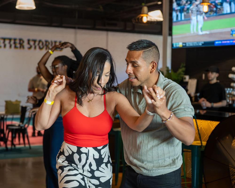 Couple dancing salsa together in a vibrant bar setting, woman in red top and floral skirt, man in sage green shirt.