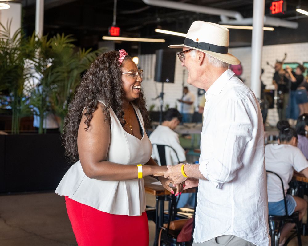 Two people shaking hands and smiling at an indoor event, woman in red skirt and man in white hat.