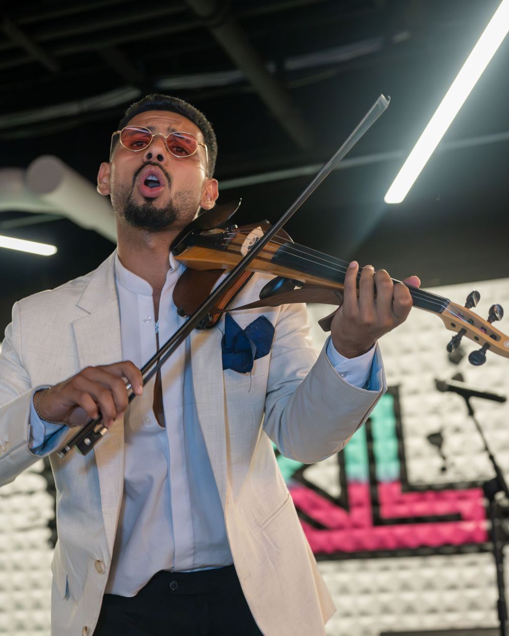 A violinist in a white blazer and pink sunglasses performs passionately on stage under bright lights.