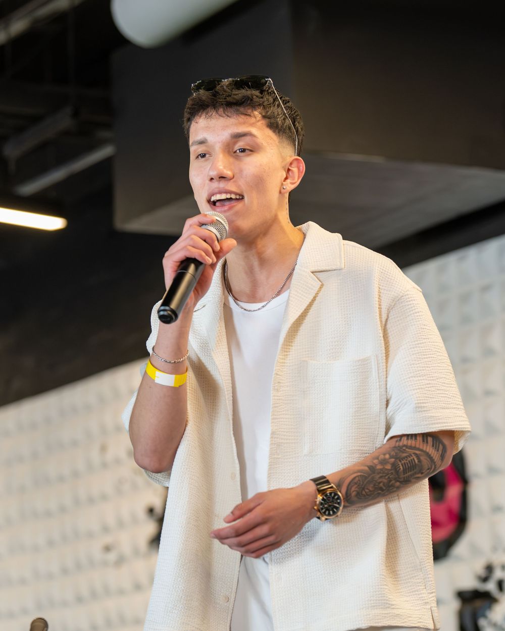 Young man with tattoos singing into a microphone, wearing a cream shirt and gold watch at an indoor event.