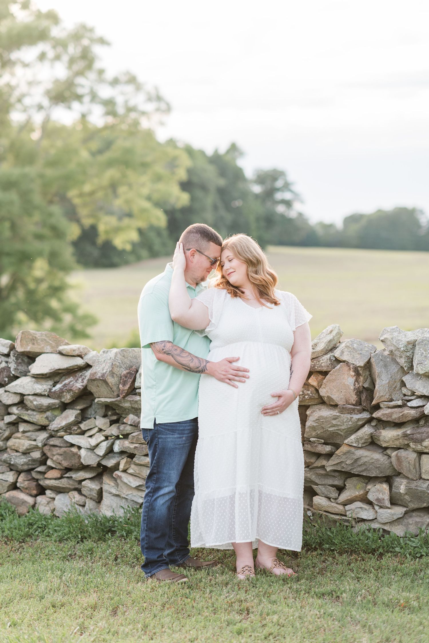 Pregnant woman in pink maxi dress posing on a porch with white columns and blooming flowers.