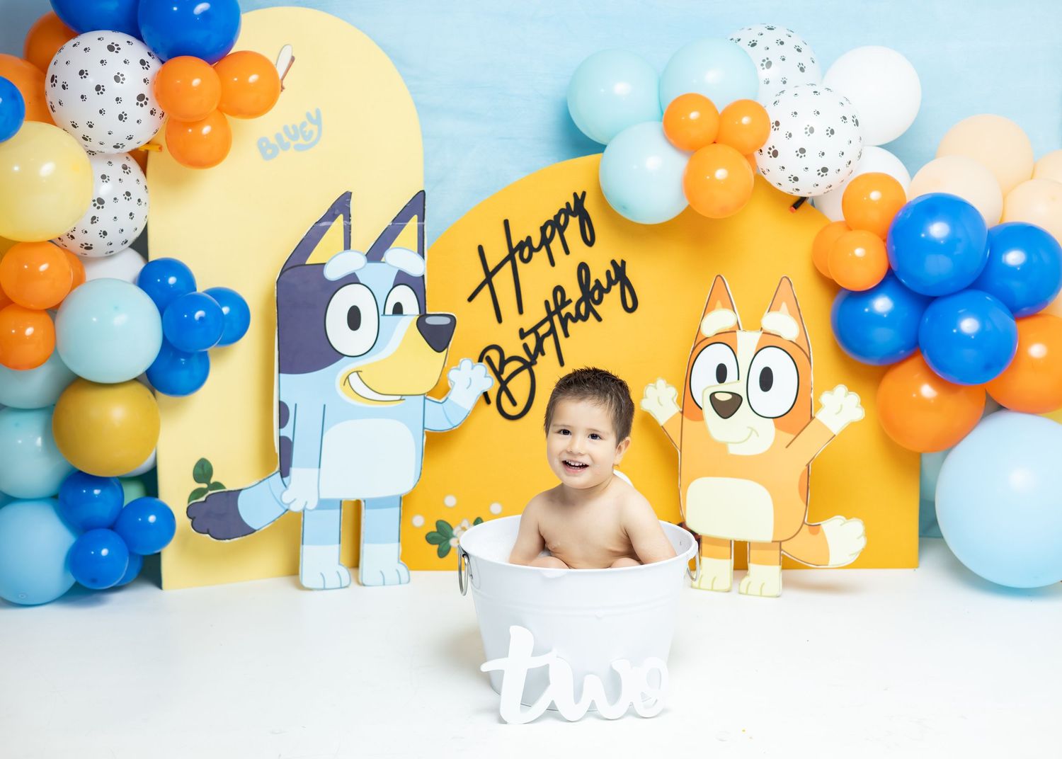Baby sits in a white tub at a Bluey-themed birthday party with colorful balloons and Happy Birthday backdrop.