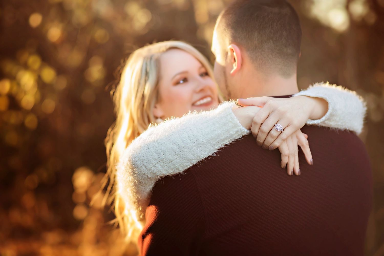 Couple embracing outdoors in autumn light, woman smiling with engagement ring visible, warm bokeh background.