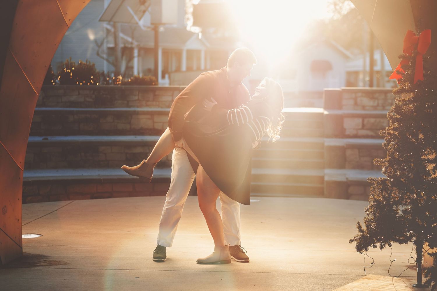 Couple sitting outdoors with pug dog holding engagement sign among autumn leaves in golden hour light.