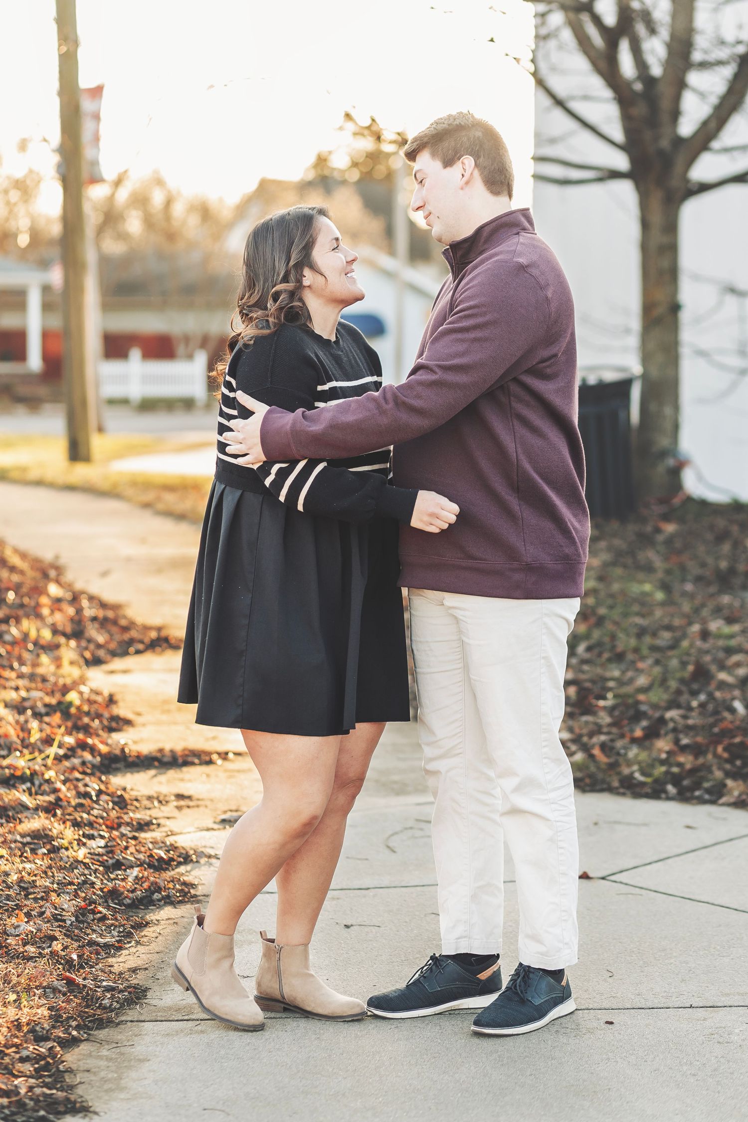 Couple embracing outdoors in autumn light, woman smiling with engagement ring visible, warm bokeh background.