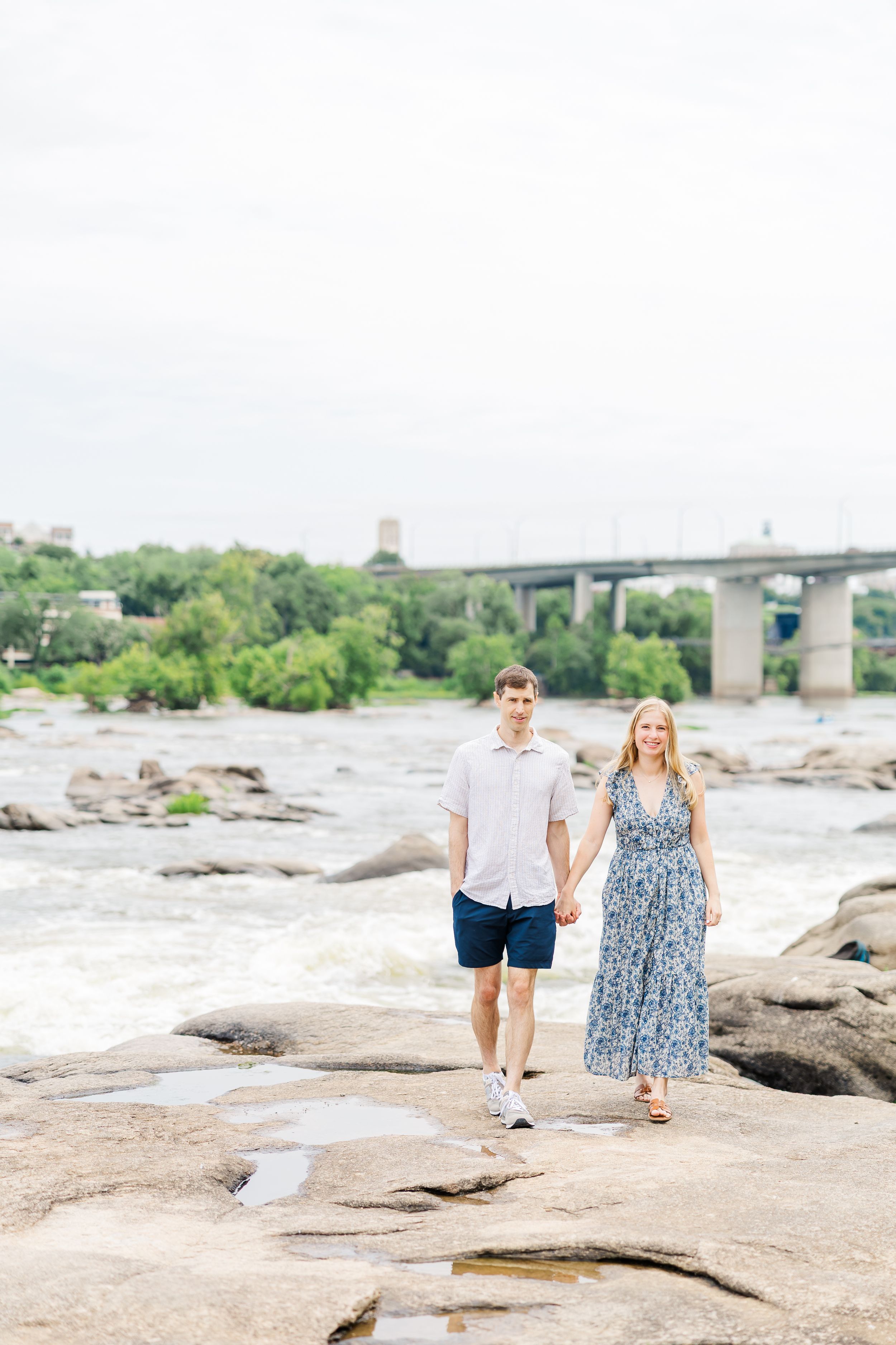 Couple holding hands walking on rocks by a river with a bridge and trees in the background.