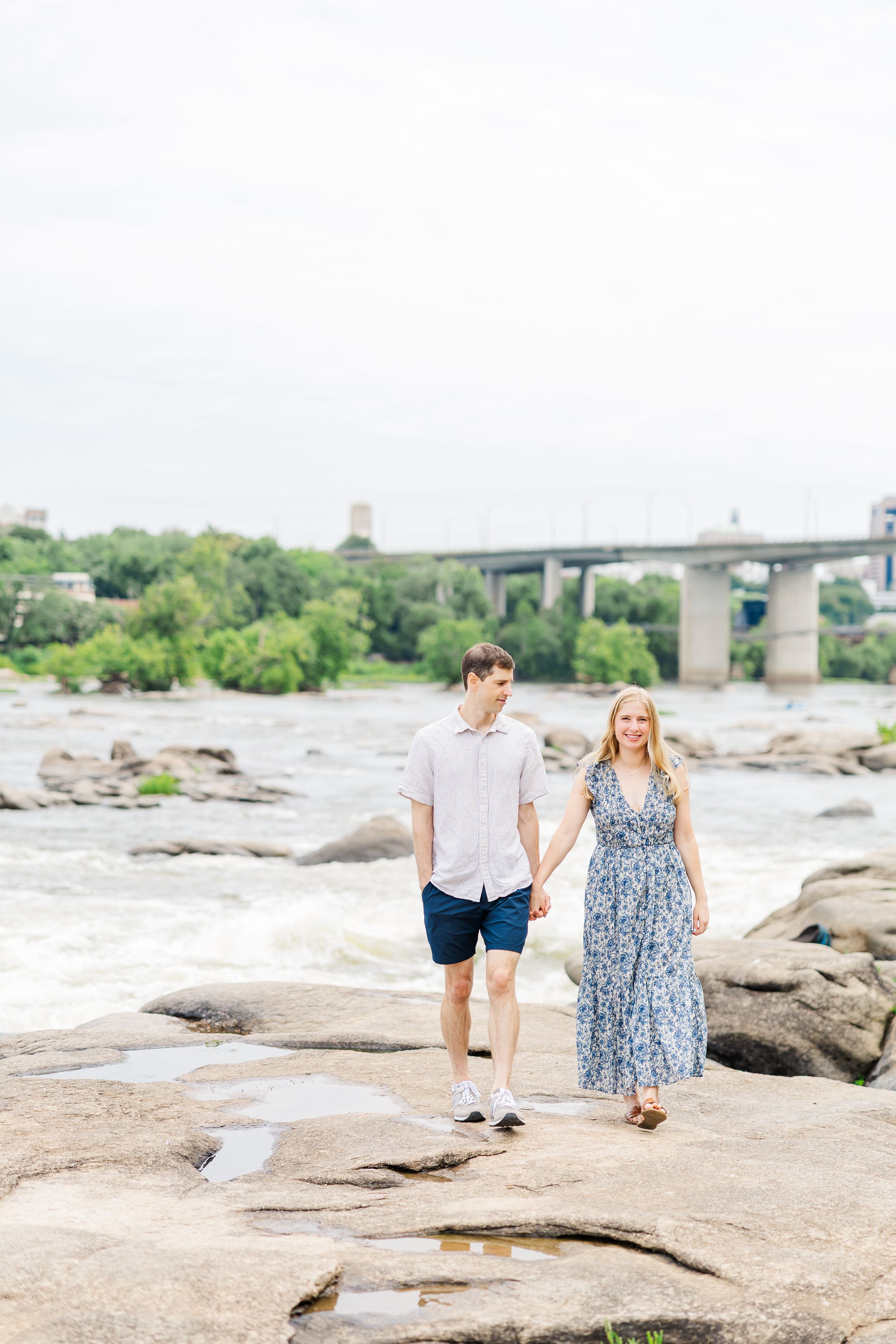 Couple holding hands on rocky riverbank with bridge and trees in background.