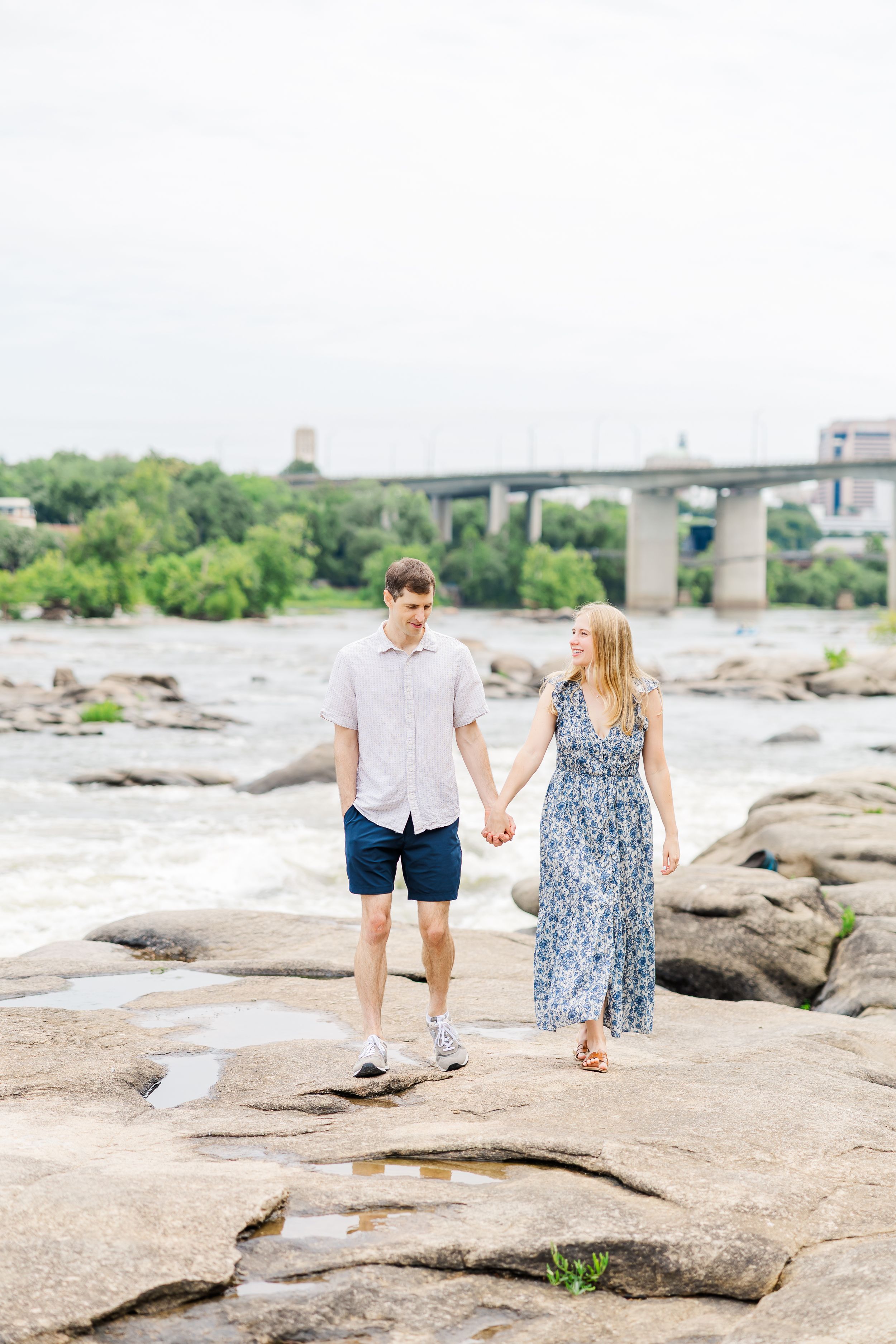 Couple holding hands walking on rocks by a river with a bridge in the background on a cloudy day.