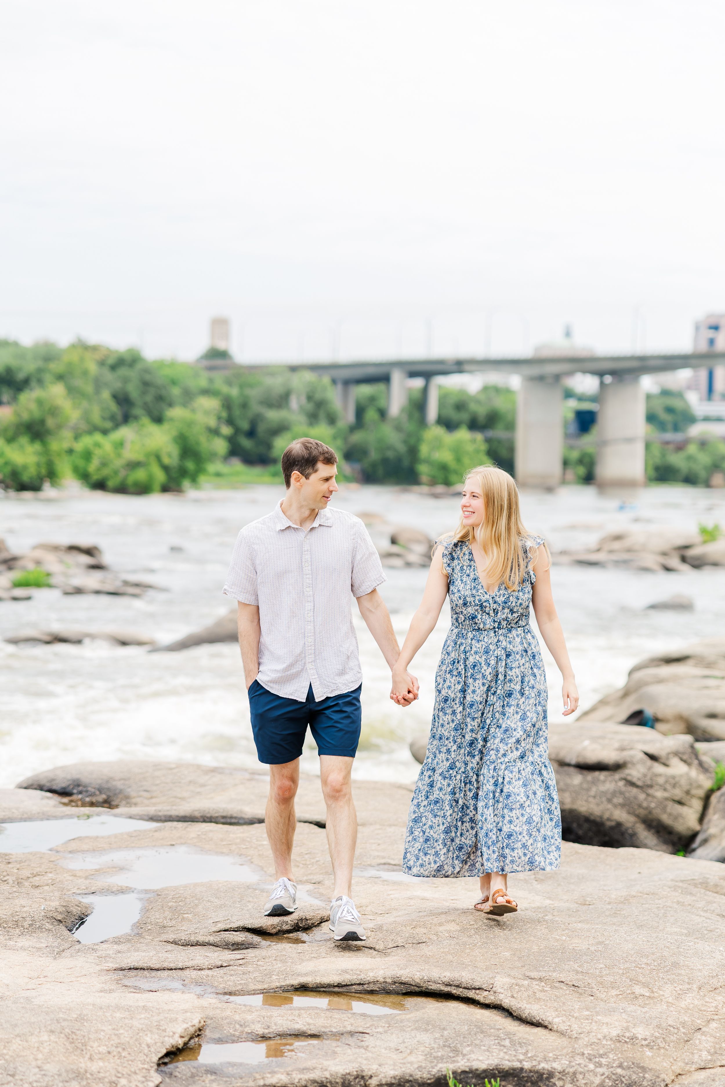 Couple holding hands on rocky riverbank with bridge and greenery in background.