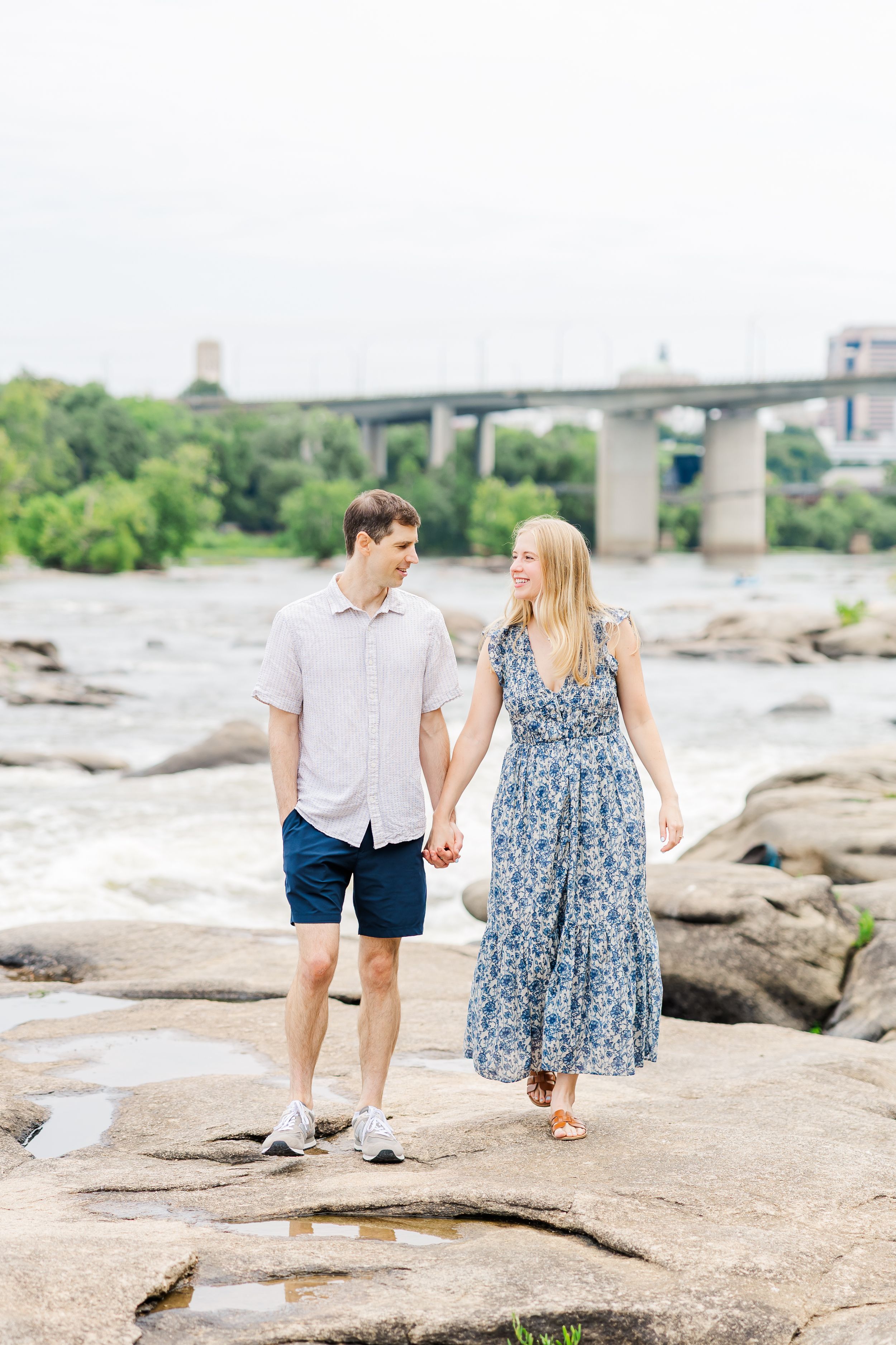 Couple holding hands walking on rocks by a river with a bridge in the background on a cloudy day.