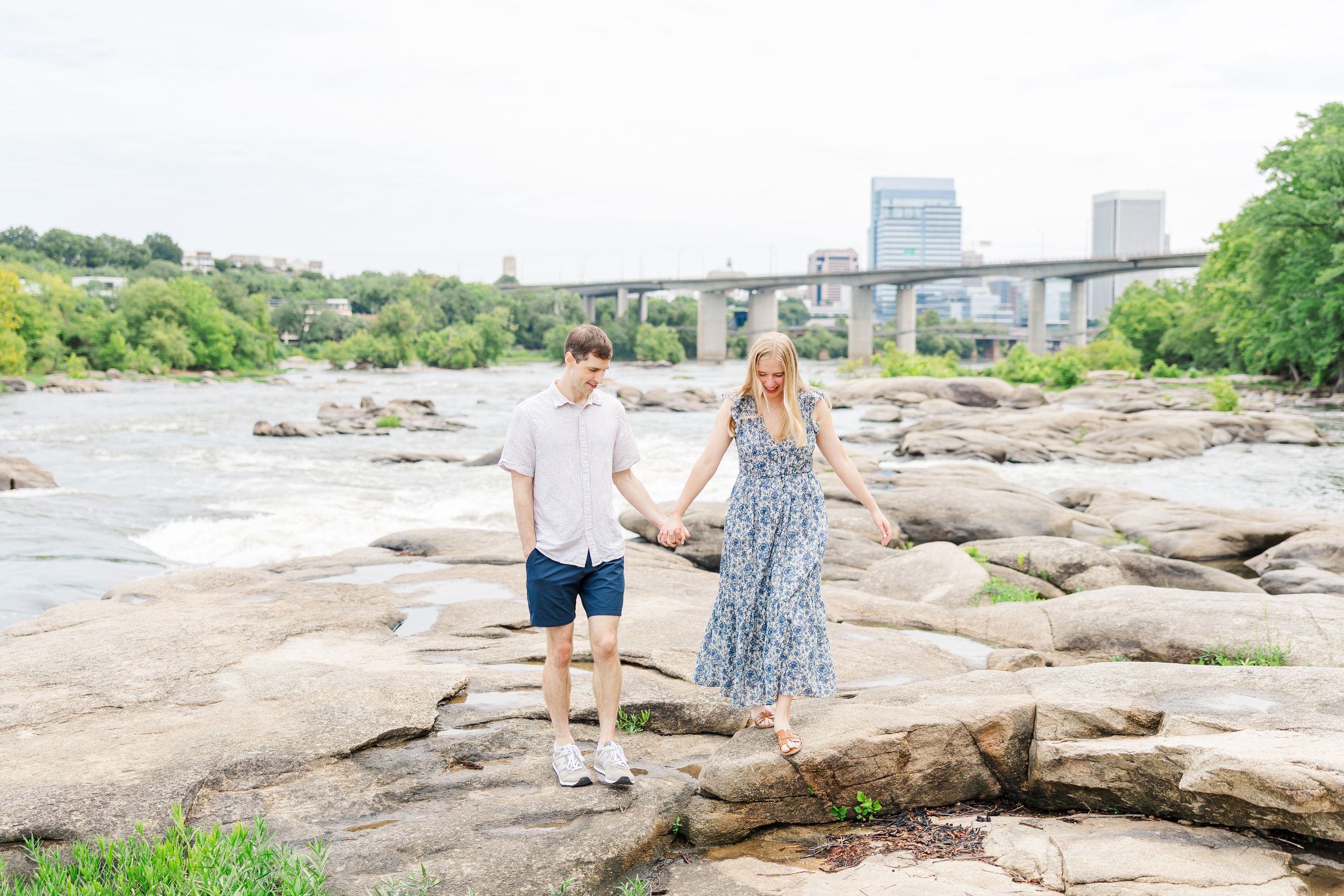 Couple holding hands on rocky riverbank with city skyline and bridge in background on a cloudy day.